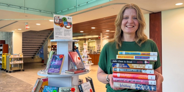 BPL chief executive officer, Lita Barrie holding a stack of books, standing beside a book display
