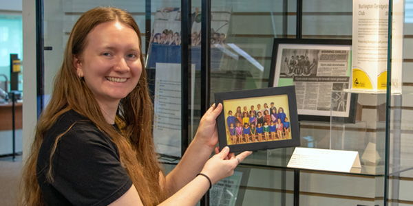BPL's Local History & Digital Archive Coordinator, Kaitlin standing at a display cabinet holding a framed photo from the Burlington Gymnastics Club collection