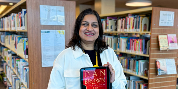 Librarian Deepti standing in front of book stacks and holding the ebook entitled The Secret of Secrets