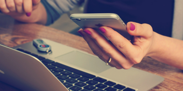 person seated at a table in fron of a laptop and holding a mobile phone