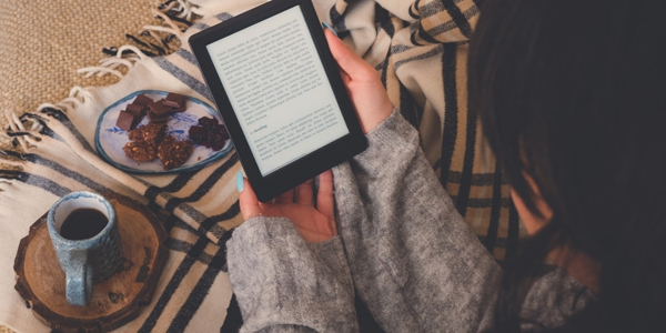 person holding an e-reader while laying on a blanket with snacks