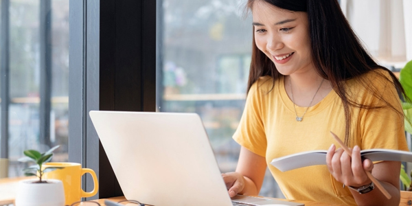 young woman seated at a table using a laptop and holding a book