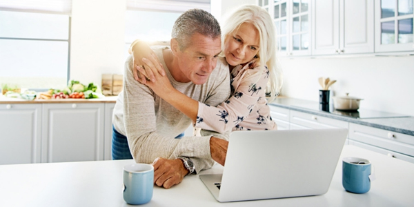 adult man and woman standing at a kitchen counter looking at a laptop with drink mugs nearby