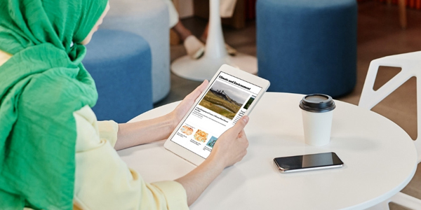 woman seated at a cafe table holding a tablet with a mobile phone and lidded takeaway cup nearby