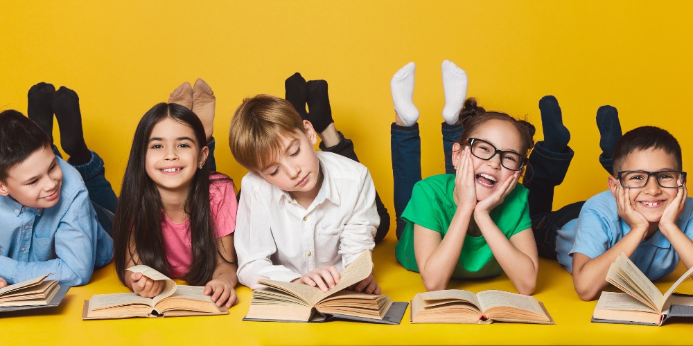 five grade-school children laying on the ground, each behind an open book.