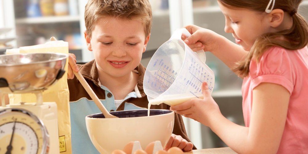 a child is pouring liquid from a measuring cup in a bowl while another child stirs