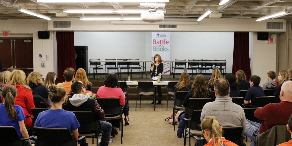 A staff member talks to students and teachers in front of a stage with two tables and twelve chairs ready for the teams.