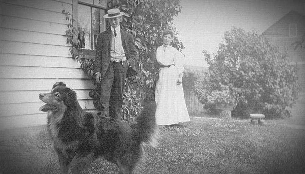 man, woman, and dog standing near a house in early 1900s