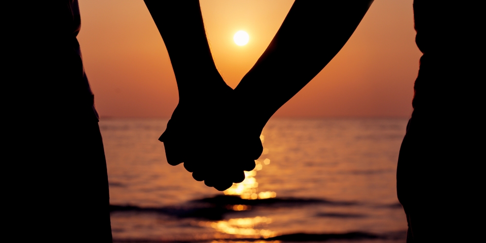 two people holding hands with the sun setting on water in the background