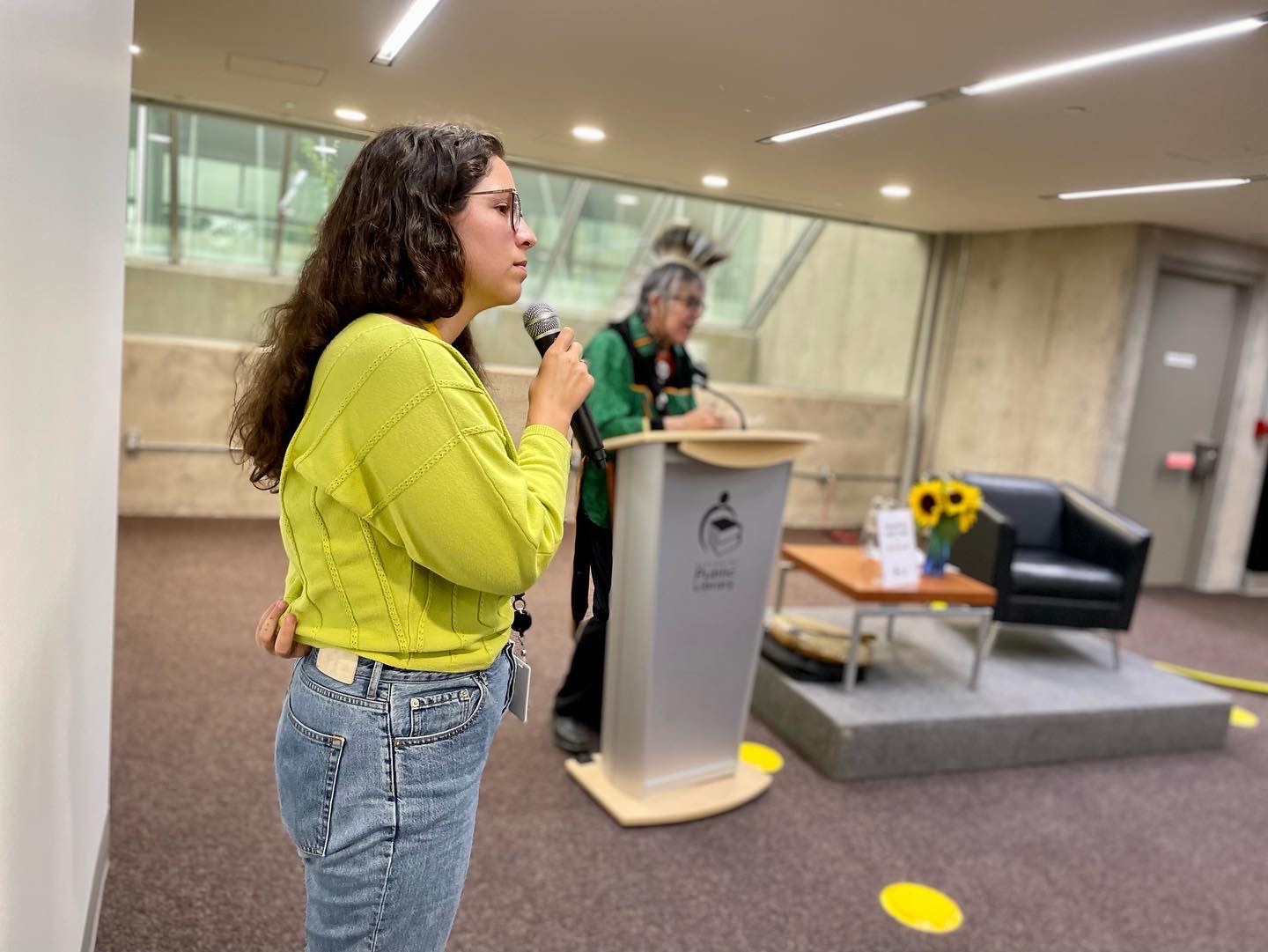 A woman in a green sweater and jeans holds a microphone. Elder Garry Sault is in the background.