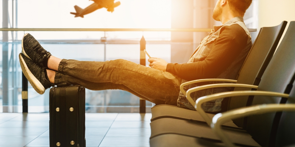 man at an airport sitting on a bank waititing chair, holding a device and resting his feet on a suitcase while looking through a window at a plane taking off