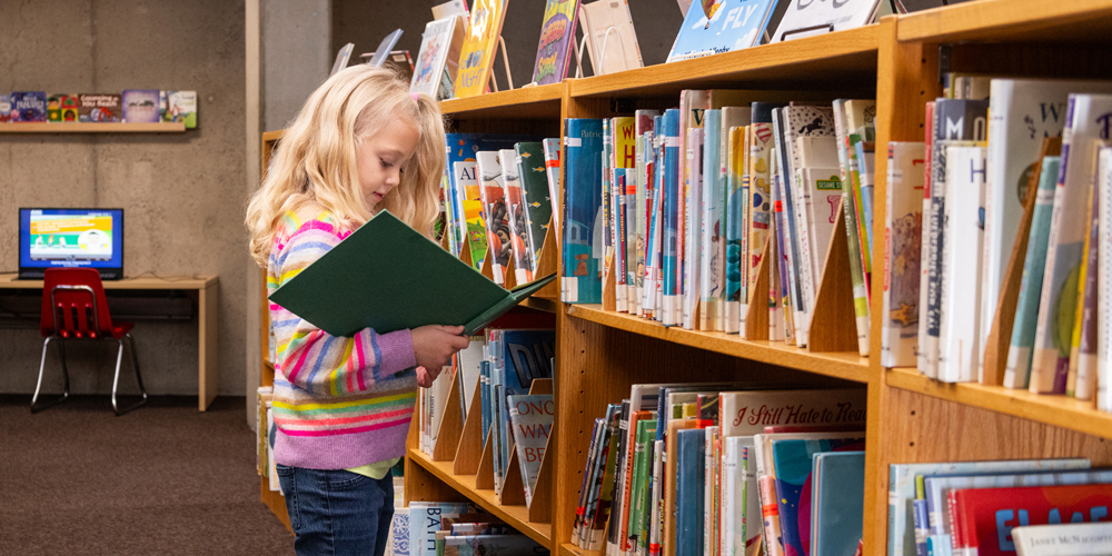 child holding an open book and standing in front of picture book shelves