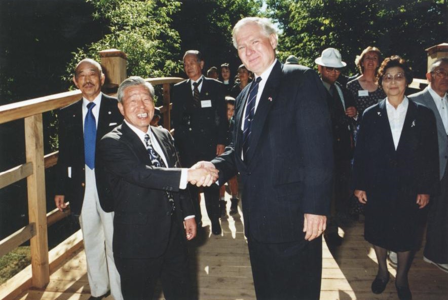 two men in suits shaking hands and smiling with a group of people standing behind