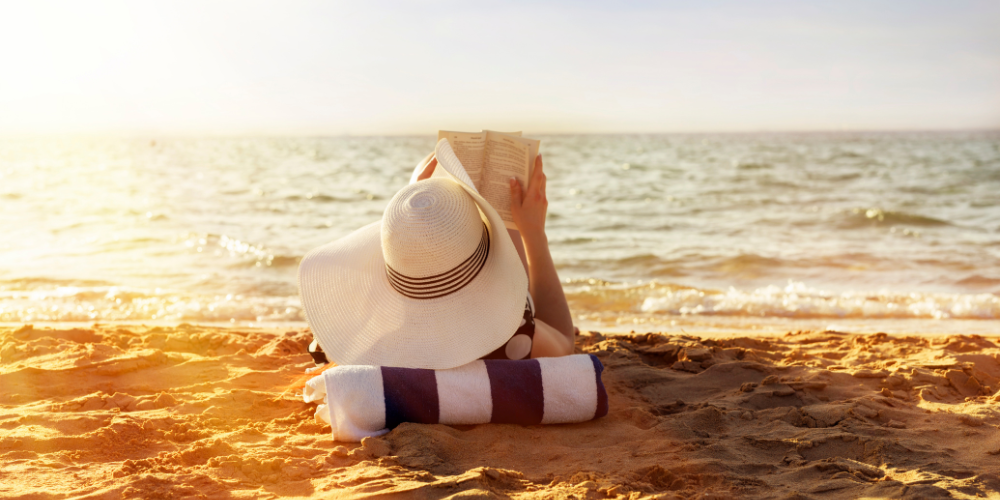 A woman lying on the beach with a large hat on and a book open in her hand