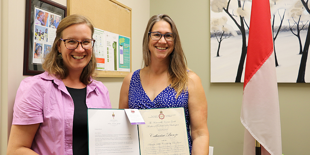 MP Karina Gould stands beside BPL Board Chair Catharine Benzie. Together they hold a large folder containing a certificate.