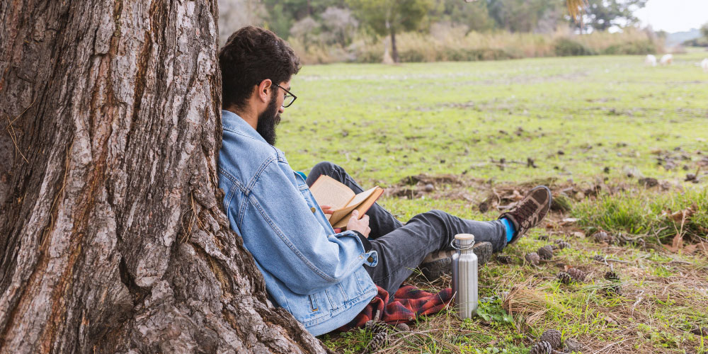 A bearded man leans up against a tree reading a book.