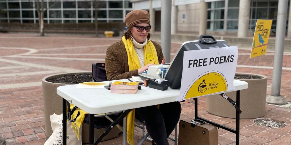 A woman in a yellow scarf sits at a desk on the sidewalk typing on an old typewriter. The typerwriter has a sign reading 'free poems' on the front