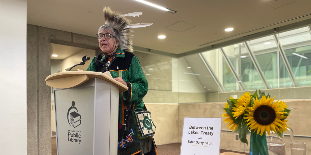 Elder Garry Sault in regalia at a podium in a large, bright and airy room.