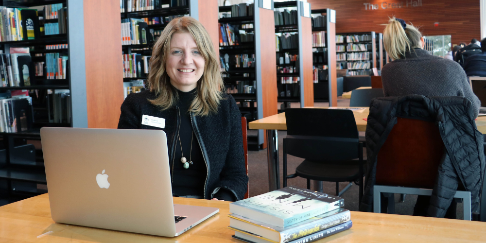 BPL CEO, Lita Barrie, sitting at a desk in front of bookshelves with a laptop open in front of her.