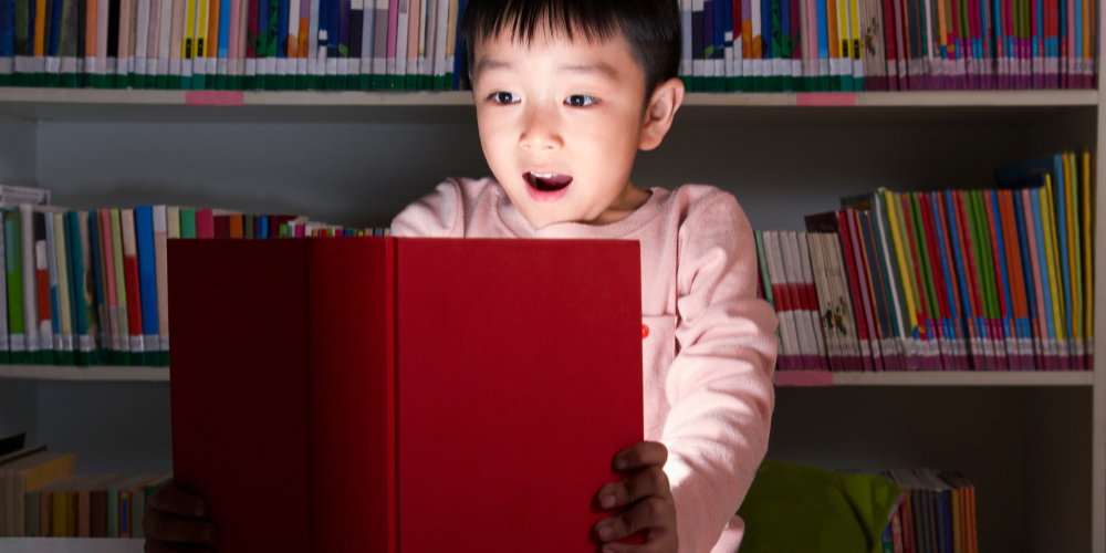 Boy opening a red book that lights up his face. A colourful shelf of books in the background.