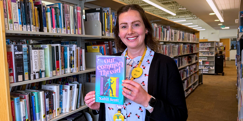 Librarian Sarah standing in front of book stacks and holding the book entitled Our Common Thread