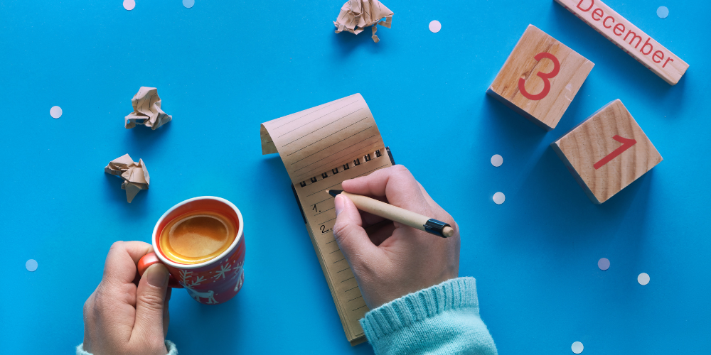 Blue background with person's hands writing a list in a notebook. Calendar blocks reading December 31 are on the surface.
