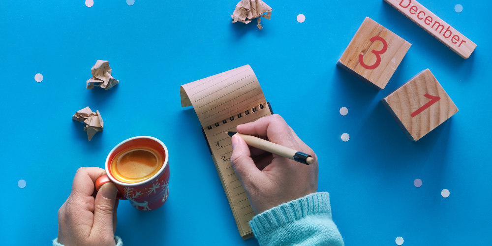 A person's hands writing a list in a notebook with calendar blocks reading December 31 on the surface of a blue table.