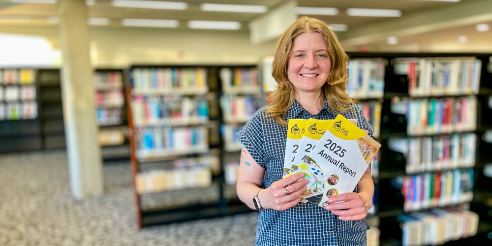 BPL CEO Lita Barrie standing near bookshelves and holding up a copy of Annual Report 2025