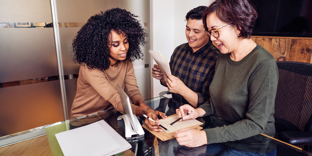 two adults sitting at a table pointing with pens to some paperwork alongside another adult holding some paper