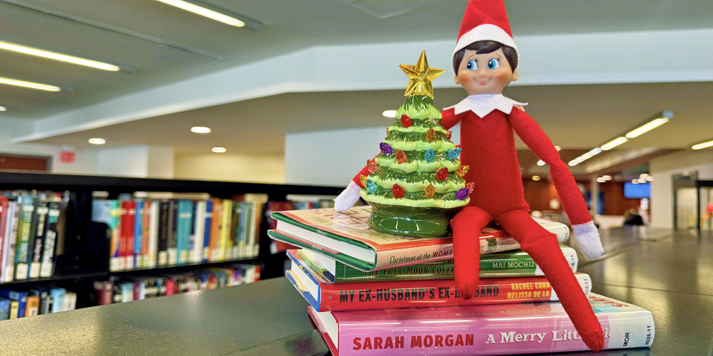 Elf on the Shelf sitting beside books and a small Christmas tree