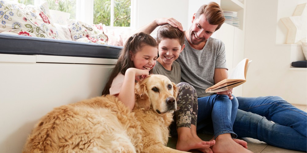 adult man holding a book, sitting on floor with dog and a laughing school-age boy and girl