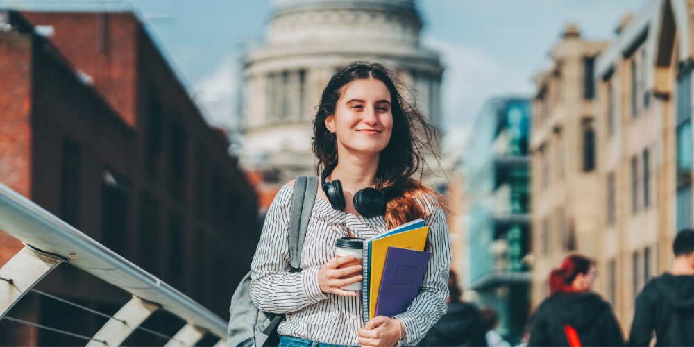 young adult holding notebooks and a coffee cup with old buildings in background