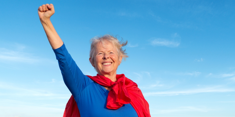 smiling older woman wearing a red cape and holding up her right fist