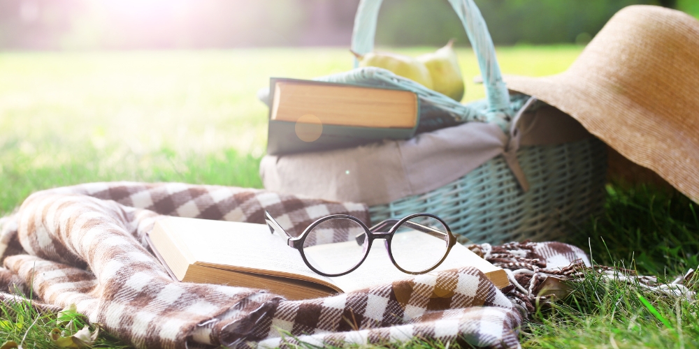 straw hat beside a picnic basket carrying a book and resting on the ground beside a checkered blanket in a green field. A book and a pair of glasses lay on the blanket.