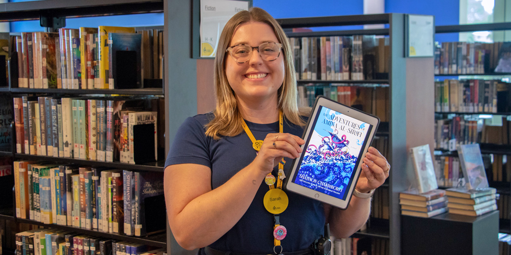 Central Branch Librarian Sarah-Ann standing in library holding The Adventures of Amina al-Sirafi