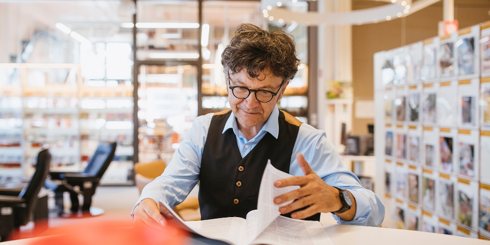 older person sitting at a table in a library turning the page of a newspaper