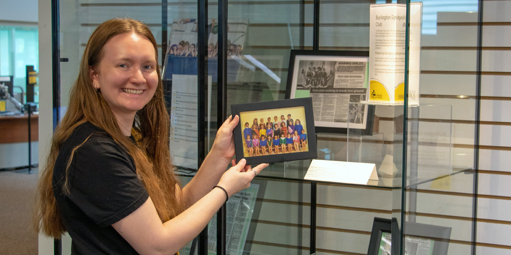 BPL's Local History & Digital Archive Coordinator, Kaitlin standing at a display cabinet holding a framed photo from the Burlington Gymnastics Club collection