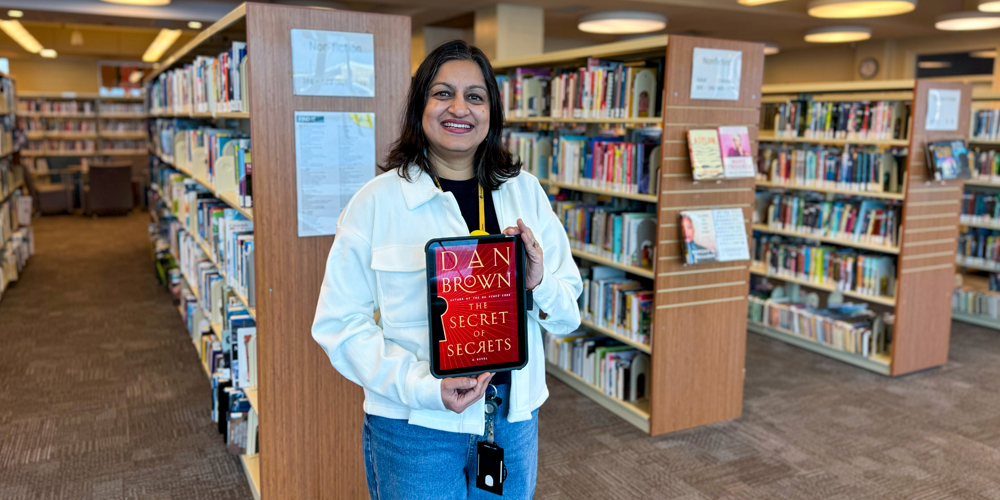 Librarian Deepti standing in front of book stacks and holding the book entitled The Secret of Secrets