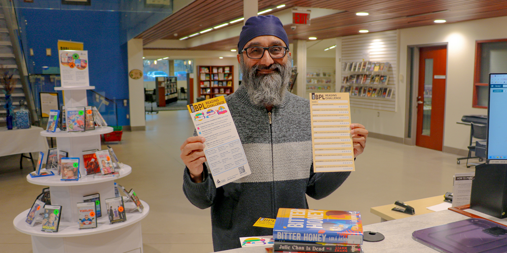 staff member holding up Reading Challenge trackers beside a pile of books