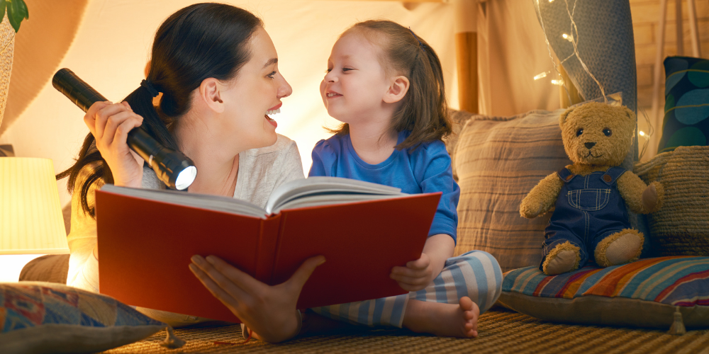 woman holding a flashlight and young girl lying on floor sharing a book with stuffed bear and pillows in background