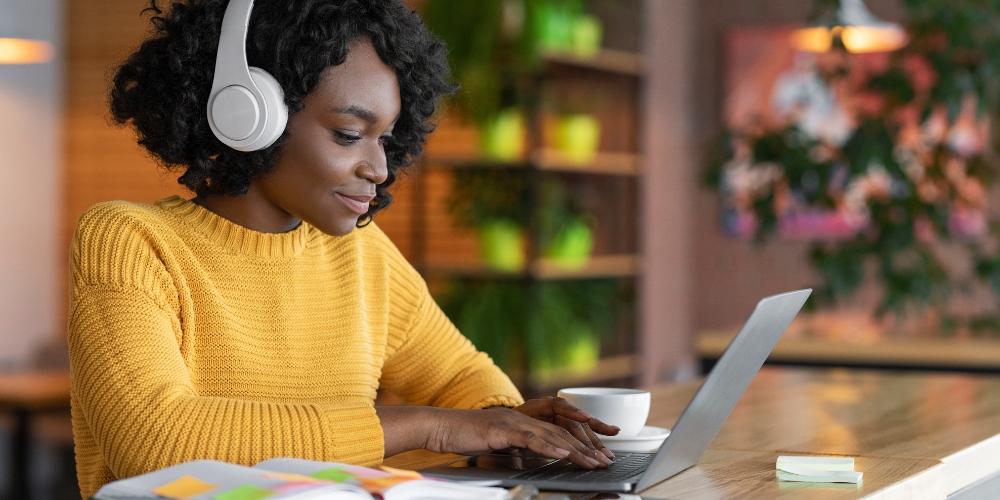 A woman at a coffee shop types on her laptop. She is wearing headphones and a bright yellow sweater. 