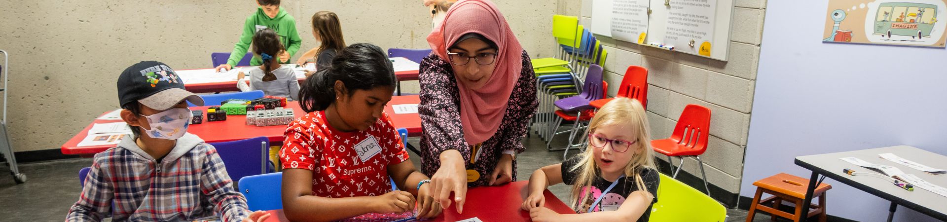 Woman in a hijab and glasses points to an activity. She is surrounded by three school-age kids at a table.